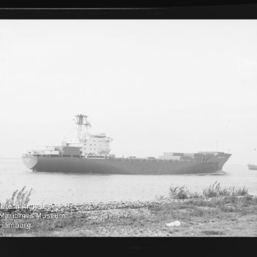 Schwarzweißfoto eines großen Frachtschiffs auf ruhigem Wasser nahe dem Ufer, mit Kiesstrand und Gras im Vordergrund, einem weiteren Schiff in der Ferne und einem Museumslogo unten links.