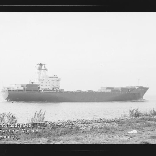 Schwarzweißaufnahme eines großen Containerschiffs in Profilsicht auf ruhiger See, davor ein grasbewachsenes Ufer; unten links sichtbar ein Wasserzeichen/Logo des Internationalen Maritimen Museums Hamburg.