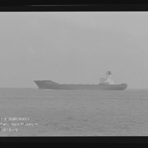 Schwarzweißaufnahme eines Frachtschiffs in mittlerer Entfernung auf ruhiger See, seitliche Silhouette mit Aufbauten nahe dem Heck vor dem Horizont, eingerahmt von Filmrand und Museumswasserzeichen.