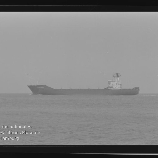 Schwarzweiß-Foto eines seitenansichtigen Frachtschiffs auf ruhiger See, mittig am Horizont, umgeben von sichtbarem Filmrahmen und mit dem Wasserzeichen ‚Internationales Maritimes Museum Hamburg‘ in der linken unteren Ecke.