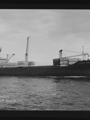 Schwarzweiß-Aufnahme eines Containerschiffs, das mit mehreren auf dem Deck gestapelten Containern und zwei Ladekränen über ruhige See fährt vor flachem Himmel; links unten sichtbar das Wasserzeichen des Internationalen Maritimen Museums Hamburg.