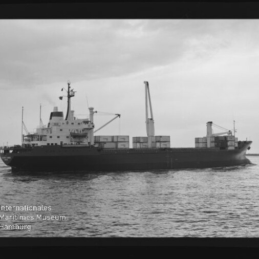 Schwarzweißaufnahme eines Containerschiffs mit gestapelten Containerkästen und einem Ladekran, das bei ruhiger See unter bewölktem Himmel von links nach rechts fährt; unten links sichtbar Logo und Schriftzug des Internationalen Maritimen Museums Hamburg.