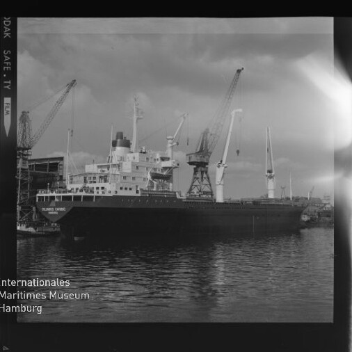 Schwarzweißaufnahme eines vor einem Werftgelände liegenden Frachtschiffs mit mehreren hohen Hafenkränen an Deck und der Spiegelung des Schiffes im ruhigen Wasser, links unten ein Museums-Wasserzeichen.