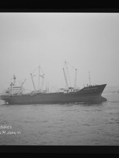 Schwarz-weiß-Foto eines Frachtschiffs mit mehreren Deckskränen vor nebligem Hafen, gerahmt wie ein Fotonegativ und mit Wasserzeichen des Internationalen Maritimen Museums Hamburg unten links.