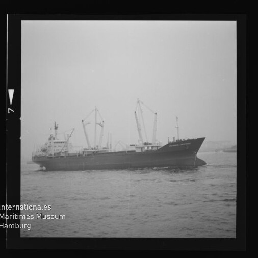 Schwarz-weiß-Foto eines Frachtschiffs mit mehreren Deckskränen vor nebligem Hafen, gerahmt wie ein Fotonegativ und mit Wasserzeichen des Internationalen Maritimen Museums Hamburg unten links.