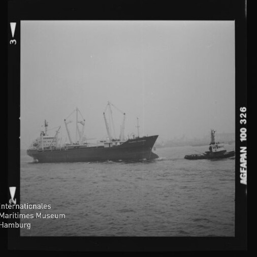 Schwarzweiß-Aufnahme eines Frachtschiffs mit mehreren Ladekränen, das auf dem Wasser von einem Schlepper begleitet wird; im Hintergrund ein nebliger Hafen und am Bildrand Filmkanten sowie das Logo des Internationalen Maritimen Museums Hamburg.