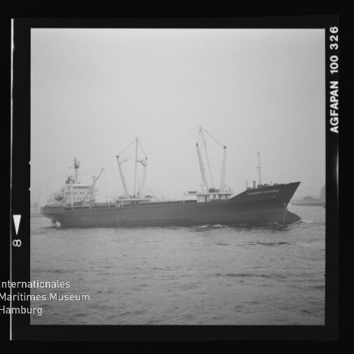 Schwarzweißaufnahme eines Frachtschiffs mit mehreren Ladekränen, das auf ruhigem Wasser fährt; im Bild sichtbar sind Filmrand mit der Aufschrift 'AGFAPAN 100 326' und das Logo sowie der Schriftzug des Internationalen Maritimen Museums Hamburg.