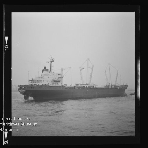 Schwarzweiß-Foto eines Frachtschiffs in Seitenansicht auf ruhiger See mit drei aufgerichteten Ladekränen, leicht nebligem Horizont sowie Museum-Logo und Negativrahmenrand sichtbar.
