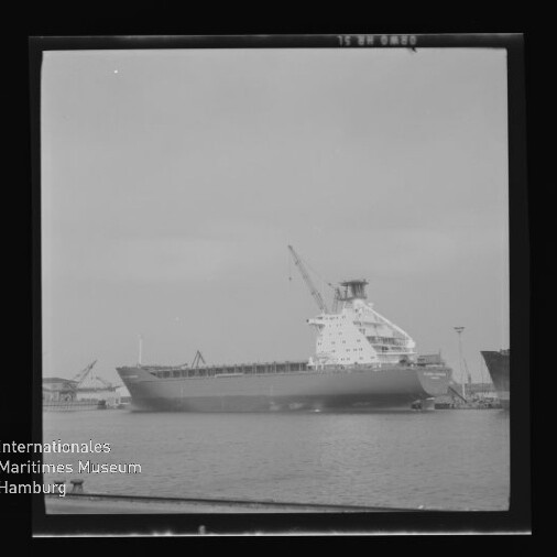 Schwarzweiß-Foto eines großen Frachtschiffs an einem Hafenpier mit aufragendem Aufbau und Kran über ruhiger Wasserfläche, eingefasst in einen quadratischen Negativrahmen; links unten Logo des Internationalen Maritimen Museums Hamburg.