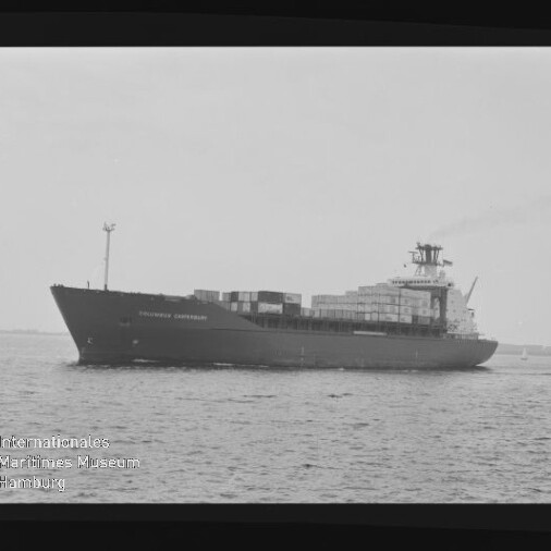 Schwarzweiß-Aufnahme eines großen Containerschiffs in Seitenansicht auf ruhiger See mit gestapelten Containercontainern, Aufbauten und leichtem Rauch aus dem Schornstein; Filmrahmen und Museumswasserzeichen unten links sind sichtbar.