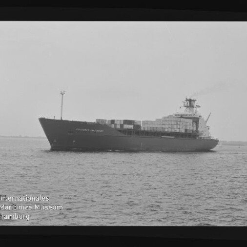Schwarzweiß-Foto eines Containerschiffs in Seitenansicht mit übereinander gestapelten Containerkästen auf dem Deck, fahrend auf ruhiger See, eingefasst von einem Filmrand und mit dem Logo des Internationalen Maritimen Museums Hamburg unten links.