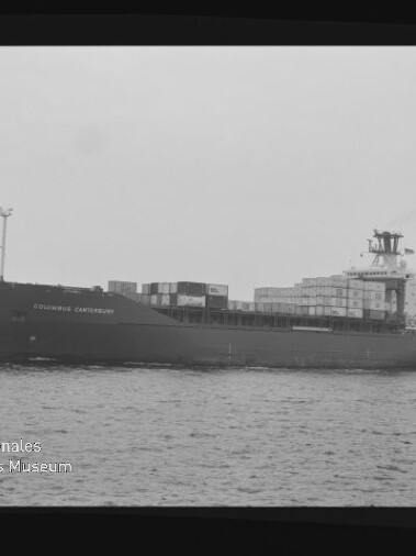 Schwarzweiß-Seitenaufnahme eines beladenen Containerschiffs mit aufgetürmten Containern auf ruhiger See; am Bug ist der Schriftzug „COLUMBUS CANTERBURY“ sichtbar und unten links ein Wasserzeichen des Internationalen Maritimen Museums Hamburg.