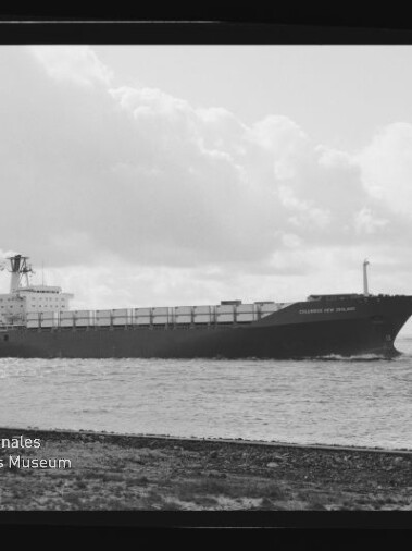 Schwarz-weiße Aufnahme eines langen Containerschiffs in Seitenansicht, das nahe der Küste auf ruhiger See unter einem wolkenverhangenen Himmel fährt.