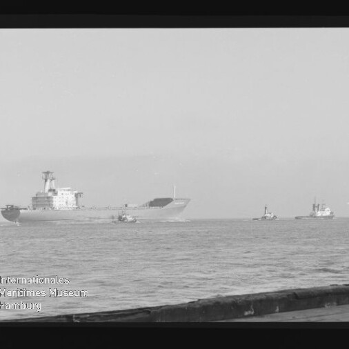 Schwarzweißfoto eines großen Frachtschiffs auf ruhiger See, begleitet von mehreren kleinen Schleppern, mit niedrigem Horizont; im Vordergrund ein Pierrand und am Bildrand sichtbare Filmränder, Beschriftung (ISOPAN 21 ISS) sowie Wasserzeichen des Internationalen Maritimen Museums Hamburg.