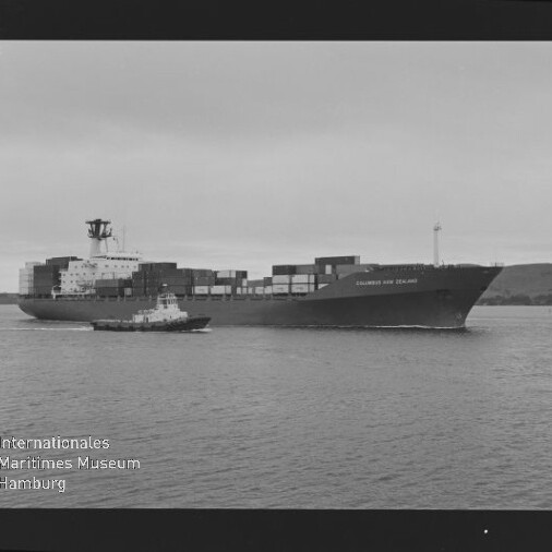 Schwarz-weiß-Foto eines beladenen Containerschiffs „Columbus New Zealand“ auf ruhigem Wasser, begleitet von einem Schlepper, mit gestapelten Containern, niedrigem Horizont, sichtbarem Filmrand (Kodak Safety Film) und Museumswasserzeichen.