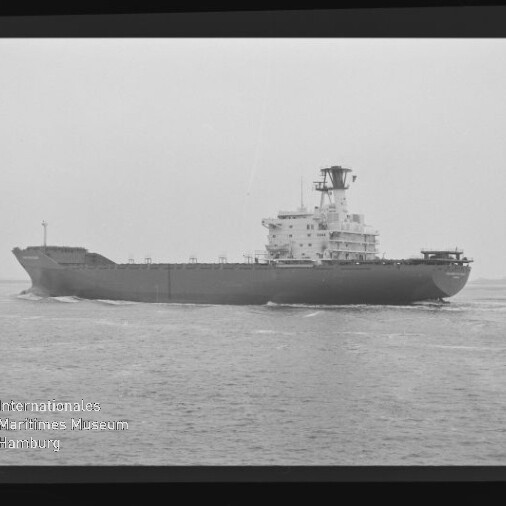 Schwarz‑weiß-Foto eines großen Frachtschiffs auf ruhiger See, seitliche Aufnahme mit hohem Brückenaufbau und Mast; Filmrand und Wasserzeichen des Internationalen Maritimen Museums Hamburg links unten sichtbar.
