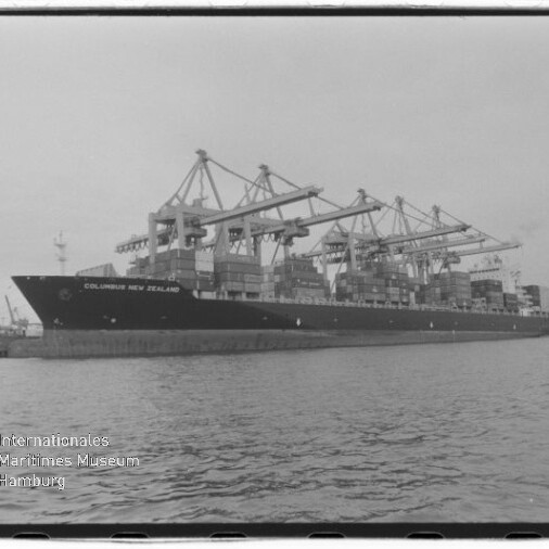 Schwarzweißaufnahme eines großen Containerschiffs 'COLUMBUS NEW ZEALAND' am Kai, beladen mit gestapelten Containern und umgeben von mehreren großen Hafenkränen, Wasser im Vordergrund und Museumslogo unten links.