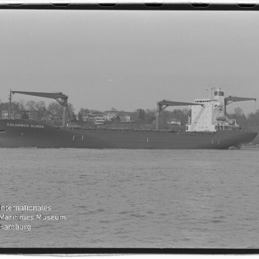 Schwarzweiß-Foto eines Frachtschiffs „COLUMBUS OLINDA“ mit zwei Ladekränen auf dem Wasser vor einem Ufer mit Häusern und Bäumen, unten links das Logo des Internationalen Maritimen Museums Hamburg sichtbar.
