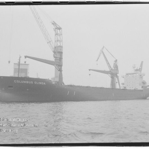 Schwarzweiß-Foto eines Frachtschiffs mit der Aufschrift COLUMBUS OLINDA, am Kai mit zwei großen Ladekränen, ruhigem Wasser im Vordergrund und Wasserzeichen des Internationalen Maritimen Museums Hamburg unten links.