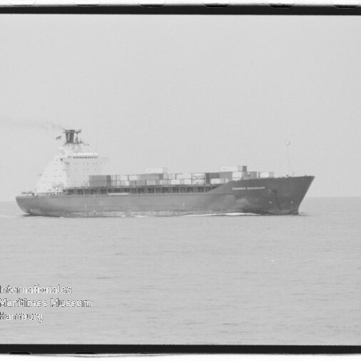 Schwarzweiß-Foto eines großen Containerschiffs in Seitenansicht auf ruhiger See mit gestapelten Containern an Deck, leichtem Rauch aus dem Schornstein und Wasserzeichen des Internationalen Maritimen Museums Hamburg unten links.