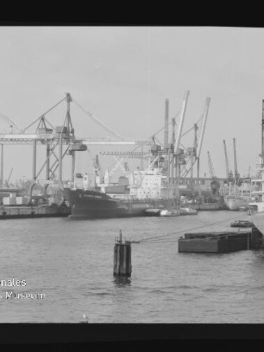 Schwarzweißaufnahme eines Industriehafens mit mehreren Frachtschiffen am Kai, zahlreichen Hafenkränen im Hintergrund, einem Dalben und einer kleinen Anlegestelle im Vordergrund sowie dem Logo des Internationalen Maritimen Museums Hamburg und sichtbarem Filmrand.