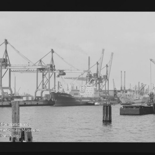 Schwarzweißaufnahme eines Industriehafens mit mehreren großen Kranen am Kai, einem festgemachten Frachtschiff in der Bildmitte, Hafenanlagen und Pollern im Vordergrund sowie dem Wasserzeichen ‚Internationales Maritimes Museum Hamburg‘ unten links.