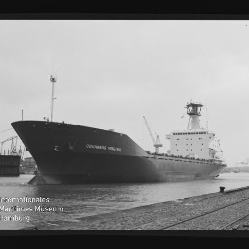 Schwarzweißaufnahme des Frachtschiffs Columbus Virginia, das an einem Industriehafen mit Kränen im Hintergrund am Kai liegt; im Vordergrund Schienen und ein Poller, unten links ein Museumswasserzeichen.
