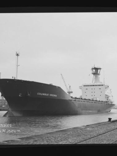 Schwarzweißaufnahme des Frachtschiffs COLUMBUS VIRGINIA am Industriehafen; im Vordergrund Kaimauer mit Pollern und Schienen, im Hintergrund Hafenkräne und -anlagen sowie ein Museumswasserzeichen.