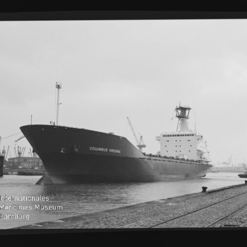 Schwarzweißaufnahme des Frachtschiffs COLUMBUS VIRGINIA am Industriehafen; im Vordergrund Kaimauer mit Pollern und Schienen, im Hintergrund Hafenkräne und -anlagen sowie ein Museumswasserzeichen.