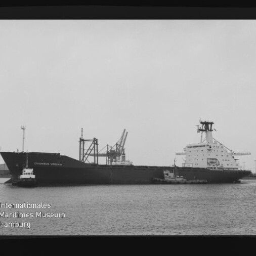 Schwarzweiß-Aufnahme eines Frachtschiffs Columbus Virginia im Hafen, seitliche Ansicht mit zwei Schleppern am Bug, Hafenkränen im Hintergrund und Wasserzeichen des Internationalen Maritimen Museums Hamburg links unten.