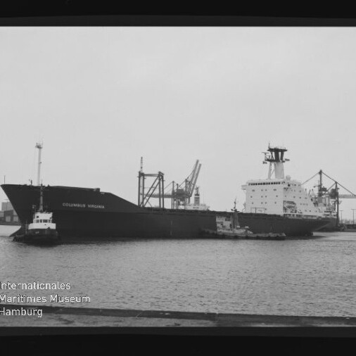 Schwarzweißaufnahme eines großen Frachtschiffs namens „Columbus Virginia“ im Hafen, von einem Schlepper begleitet, vor Kaianlagen und Ladekränen; Museumslogo unten links.