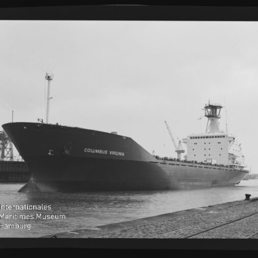 Großes Frachtschiff „COLUMBUS VIRGINIA“ in Schwarz-Weiß, am Kai eines Industriehafens festgemacht mit sichtbarem Schiffsrumpf, Kranen im Hintergrund und Wasserzeichen des Internationalen Maritimen Museums Hamburg.