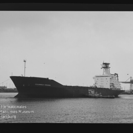 Schwarzweißfoto eines großen Frachtschiffs COLUMBUS VIRGINIA im Hafen, ein Schlepper am Heck, Hafenkräne im Hintergrund, Negativrahmen mit Nummer links und Museumssignet unten links.