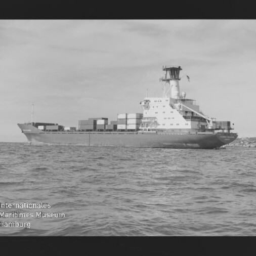 Schwarzweiß-Foto einer Seitenansicht eines Containerschiffs mit gestapelten Containern und markantem Brückenaufbau, das auf ruhiger See vor einer flachen Küstenlinie fährt; sichtbarer Filmrand und Wasserzeichen 'Internationales Maritimes Museum Hamburg' unten links.