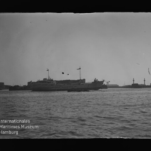 Schwarzweiß-Foto eines großen Passagier- oder Fährschiffs mit gehissten Flaggen auf ruhigem Hafenwasser, im Hintergrund Silhouetten von Landanlagen und markanten Türmen; links unten sichtbares Wasserzeichen des Internationalen Maritimen Museums Hamburg sowie altersbedingte Flecken und Kratzer im Negativ.