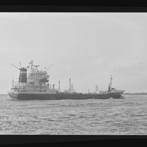 Schwarz-weiß-Foto eines Frachtschiffes/Tankers in Seitenansicht auf ruhiger See mit sichtbaren Aufbauten und Ladeeinrichtungen, Wellen im Vordergrund und dem Wasserzeichen des Internationalen Maritimen Museums Hamburg unten links.