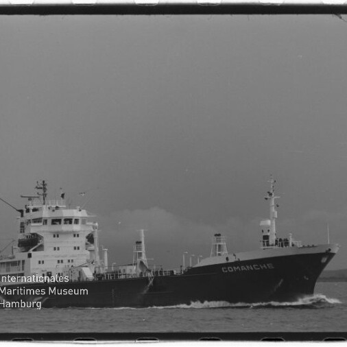 Schwarz‑weiß-Foto eines Frachtschiffs namens COMANCHE, seitliche Ansicht auf See; links unten Wasserzeichen des Internationalen Maritimen Museums Hamburg.