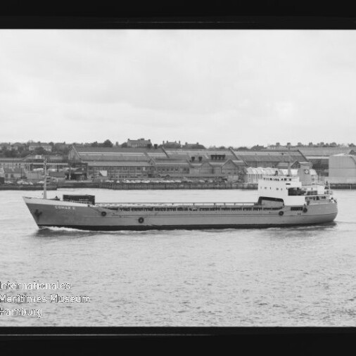 Flaches Frachtschiff fährt auf ruhigem Wasser vor einer Industriehafen-Kulisse mit Lagerhallen; links unten ist das Logo des Internationalen Maritimen Museums Hamburg zu sehen.