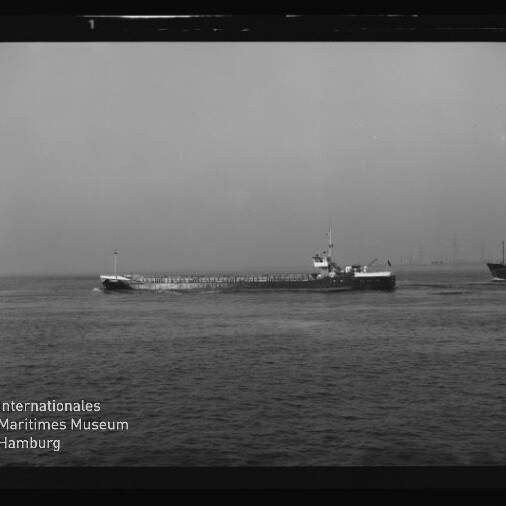 Schwarzweißaufnahme eines langen, niedrigen Frachtschiffs, das quer durchs Bild in ruhigem Wasser fährt; im Hintergrund eine ferne Küstenlinie mit Strommasten und rechts am Bildrand ein weiteres Schiff, unten links das Logo und der Schriftzug Internationales Maritimes Museum Hamburg.
