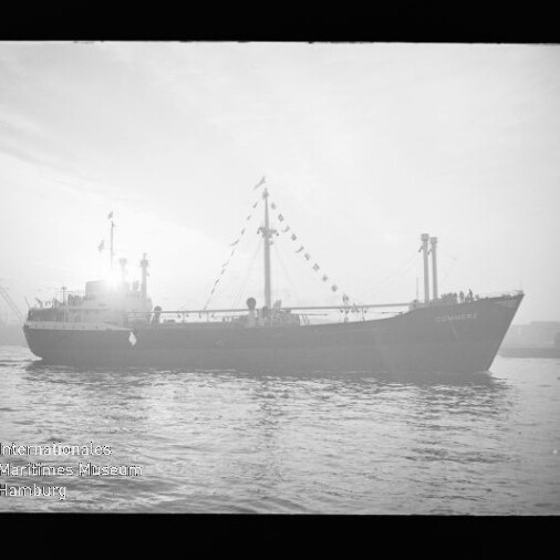 Schwarzweiß-Foto eines Frachtschiffs in Seitenansicht, silhouettenhaft vor hellem Himmel mit einer Girlande aus Signalflaggen am Mast, ruhigem Wasser im Vordergrund und Hafenkränen im Hintergrund; Wasserzeichen des Internationalen Maritimen Museums Hamburg unten links.
