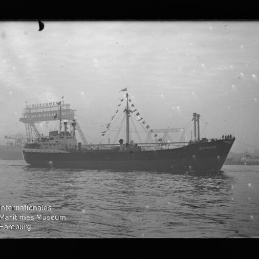 Schwarz-weiß-Foto eines beflagten Frachtschiffs in seitlicher Ansicht auf ruhiger See, mit Hafenbauten im Hintergrund und Wasserzeichen des Internationalen Maritimen Museums Hamburg.