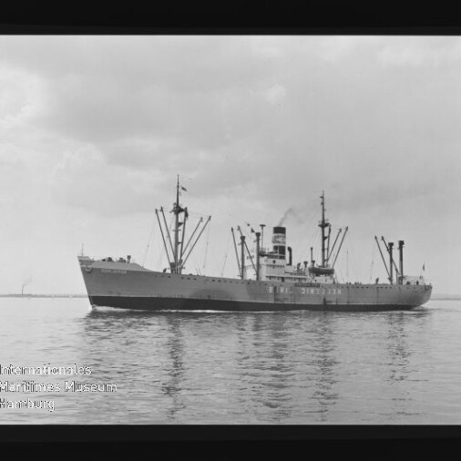 Schwarzweißaufnahme eines Frachtschiffs mit mehreren Ladebäumen und einem Schornstein, das ruhig über spiegelndes Wasser fährt; Filmrand mit KODAK-Markierung und Wasserzeichen des Internationalen Maritimen Museums Hamburg sichtbar.