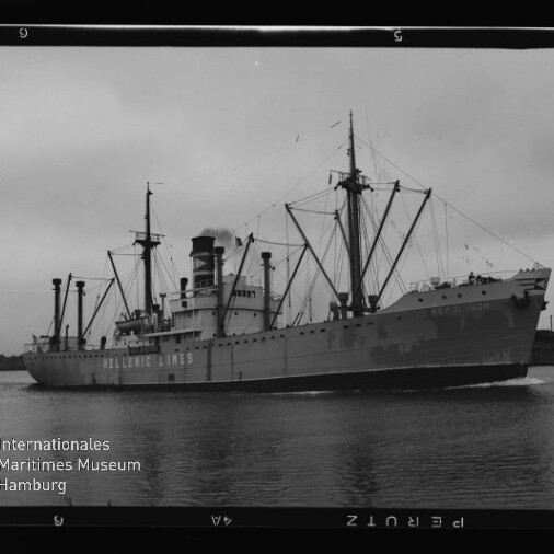 Schwarzweiß-Foto eines Hellenic Lines Frachtschiffs mit markantem Schornstein, Ladekränen und Masten, das ruhig auf dem Wasser an einer flachen Küstenlinie vorbeifährt; Filmbildrand und Wasserzeichen des Internationalen Maritimen Museums Hamburg sichtbar.