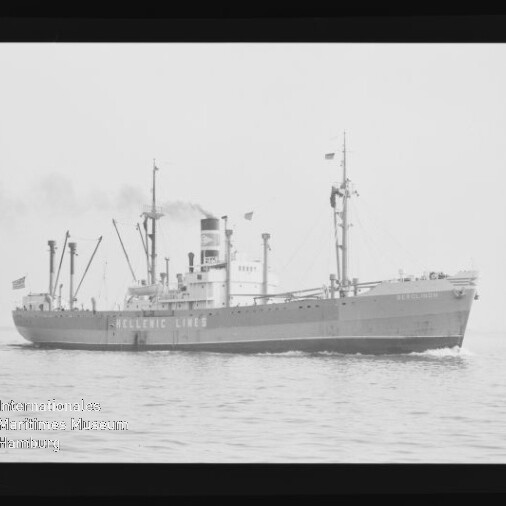 Schwarzweiß-Foto eines Frachtschiffs der Reederei Hellenic Lines mit hohem Schornstein, mehreren Ladekränen und Flaggen, das bei ruhiger See eine leichte Bugwelle zieht.