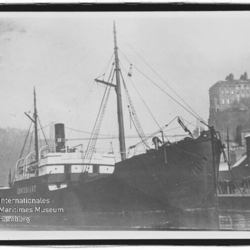 Schwarzweiß-Aufnahme eines im Hafen angelegten Dampfschiffs mit Schornstein, Masten und Ladegeschirr, rechts Kai mit Personen und Gebäuden auf einer Anhöhe; unten links sichtbar das Logo des Internationalen Maritimen Museums Hamburg.