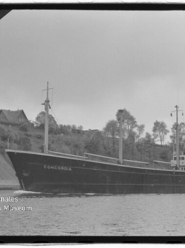 Schwarzweißaufnahme eines Frachtschiffs namens CONCORDIA, das auf einem Fluss entlang eines bewaldeten Ufers unter einem Brückenbogen fährt, im Hintergrund Häuser und Bäume.