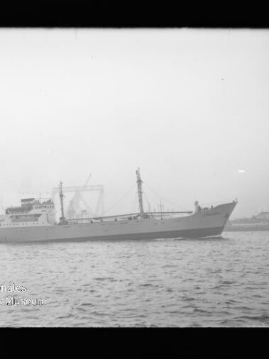 Graustufiges Foto eines Frachtschiffs auf ruhigem Wasser vor einem nebligen Hafen mit Hafenkränen und Werftgebäuden im Hintergrund; links unten sichtbares Wasserzeichen ‚Internationales Maritimes Museum Hamburg‘.