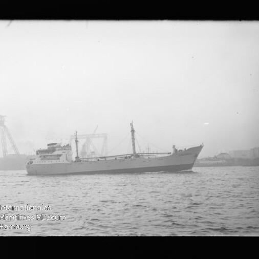 Graustufiges Foto eines Frachtschiffs auf ruhigem Wasser vor einem nebligen Hafen mit Hafenkränen und Werftgebäuden im Hintergrund; links unten sichtbares Wasserzeichen ‚Internationales Maritimes Museum Hamburg‘.
