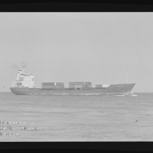 Schwarzweiß-Aufnahme eines Containerschiffs in Seitenansicht auf ruhiger See mit gestapelten Containern auf dem Deck, dem Aufbauten/Brückenturm hinten links, sichtbaren Filmrändern und einem Museumswasserzeichen unten links.