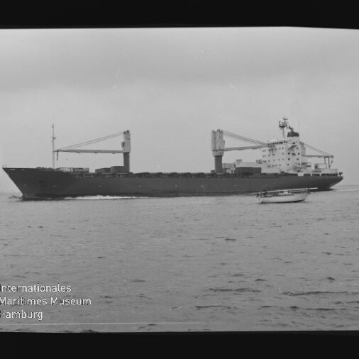 Schwarzweiß-Foto eines Frachtschiffs mit zwei Ladekränen, das auf ruhiger See fährt; im Vordergrund ein kleines Motorboot, unten links Wasserzeichen des Internationalen Maritimen Museums Hamburg und sichtbarer Filmrand.